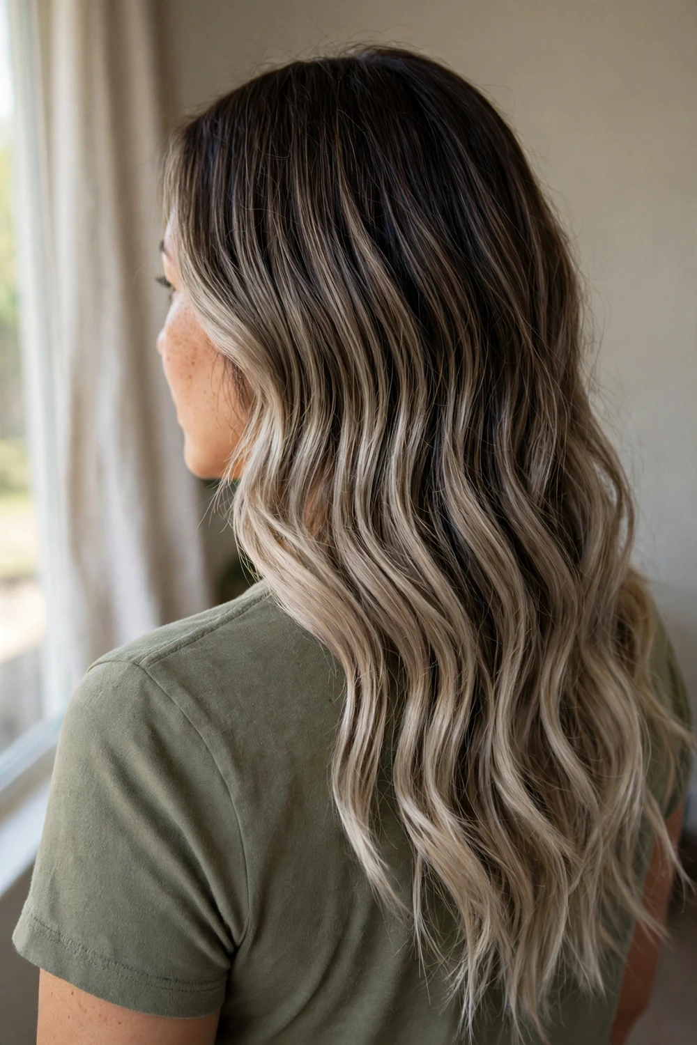 Woman with ash beige balayage in long wavy hair blending dark roots into beige-ash ends