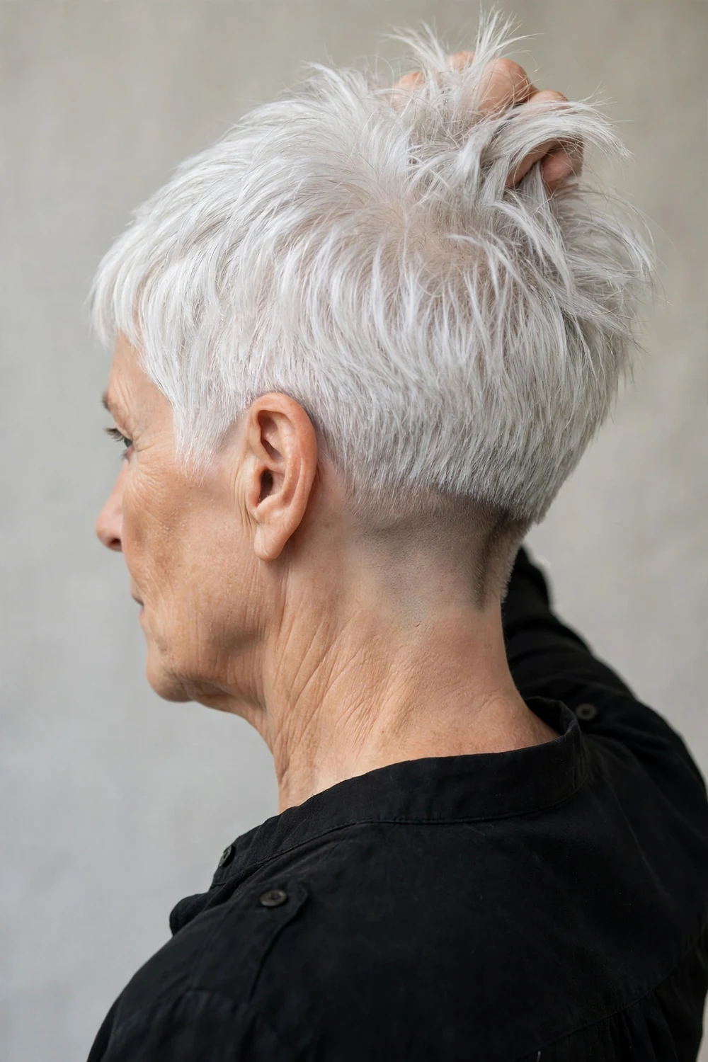 Caucasian woman in her late 60s with a white choppy pixie, top lifted to show a hidden shaved nape undercut